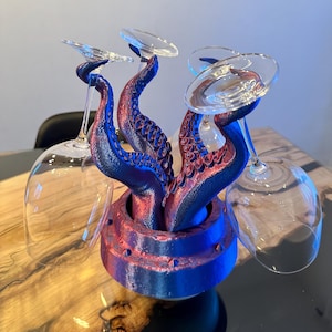 Peut inclure: Un porte-verres à vin en forme de poulpe, avec des tentacules bleues et rouges soutenant des verres transparents. Le support a une base circulaire et est posé sur une table en bois. Un design créatif pour présenter les verres à vin.