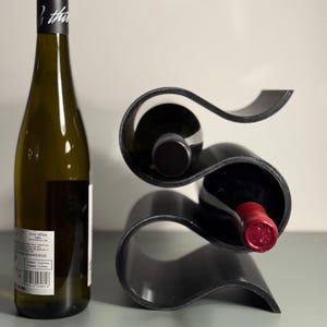 May include: A black, modern wine rack holding two bottles of wine, with a third bottle standing upright to the left. The wine rack has a unique, curved design. The wine bottle on the left has a black cap with white text.