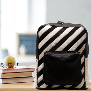 Puede incluir: Una mochila a rayas de cebra en blanco y negro con un bolsillo de cuero negro. La mochila tiene forma rectangular con cierre de cremallera. La bolsa está hecha de un material texturizado y es adecuada para llevar libros y otros útiles escolares.