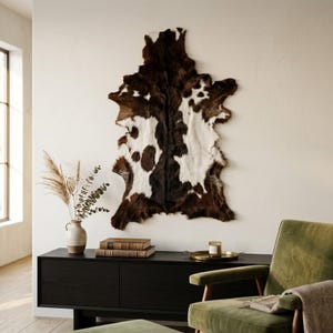 May include: A cowhide rug with a natural pattern of dark brown and white patches, displayed as a wall hanging. The hide features an irregular, organic shape with a dense, short-haired texture, showcasing a high-contrast color distribution.