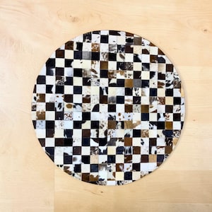 May include: A circular cowhide rug with a chequered design. The rug is made up of squares of black, white, and brown cowhide. This rug is a decorative accent piece for a living room or bedroom.