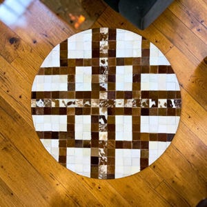 May include: A round rug with a geometric pattern. The rug features a patchwork design of white, brown, and tan squares. The rug is made of a natural material, likely cowhide, and has a soft texture. The rug is placed on a wooden floor.