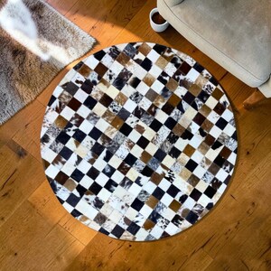 May include: A round rug with a patchwork design. The rug features a checkerboard pattern with squares of various colors and textures, including white, black, brown, and tan. The rug is made of a natural material, likely cowhide.
