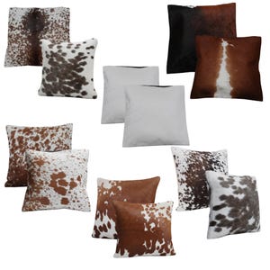 May include: A collection of square throw pillows featuring various cowhide patterns in shades of brown, black, and white, alongside solid white pillows. The textures display natural animal hide markings, including spotted, mottled, and solid color blocks.