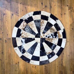 May include: A round cowhide rug with a striking checkerboard design. The rug features alternating black and white squares, with sections of natural cowhide in shades of brown, black, and white. The rug is placed on a wooden floor.