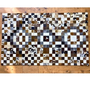 May include: A rectangular cowhide rug with a patchwork design. The rug features a checkerboard pattern of squares in various shades of brown, white, and black. The rug is made of natural cowhide material.