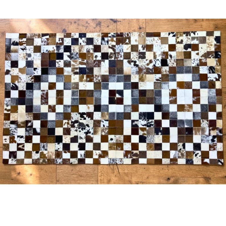 K&ouml;nnte beinhalten: Ein rechteckiger Kuhfellteppich mit Patchwork-Design. Der Teppich hat ein Schachbrettmuster aus Quadraten in verschiedenen Braun-, Wei&szlig;- und Schwarzt&ouml;nen. Der Teppich besteht aus nat&uuml;rlichem Kuhfellmaterial.