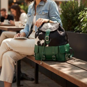 May include: A green leather backpack with a black and white cowhide flap. The backpack has two front pockets and silver-coloured buckles. The bag is on a wooden bench.