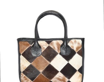 May include: A rectangular tote bag with a patchwork design. The bag features a black leather trim and handles. The patchwork is made of diamond-shaped pieces in shades of brown, tan, and white. The bag has a structured shape and a classic design.
