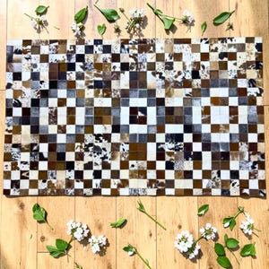 K&ouml;nnte beinhalten: Ein rechteckiger Kuhfellteppich mit einem Patchwork-Design. Der Teppich hat ein Schachbrettmuster aus Quadraten in Braun-, Wei&szlig;- und Schwarzt&ouml;nen. Der Teppich ist von wei&szlig;en Blumen und gr&uuml;nen Bl&auml;ttern auf einer Holzoberfl&auml;che umgeben.