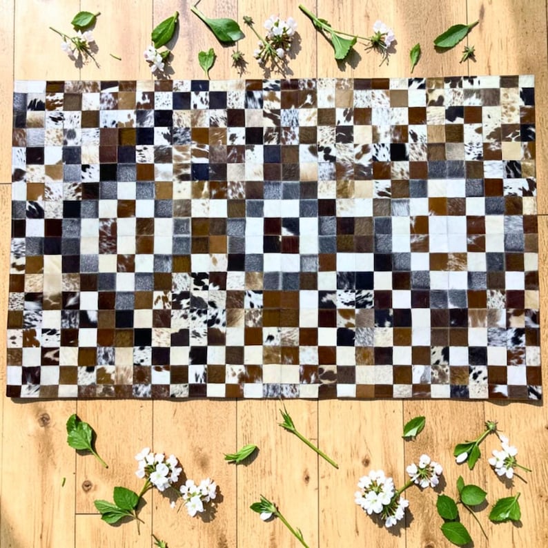 K&ouml;nnte beinhalten: Ein rechteckiger Kuhfellteppich mit einem Patchwork-Design. Der Teppich hat ein Schachbrettmuster aus Quadraten in Braun-, Wei&szlig;- und Schwarzt&ouml;nen. Der Teppich ist von wei&szlig;en Blumen und gr&uuml;nen Bl&auml;ttern auf einer Holzoberfl&auml;che umgeben.