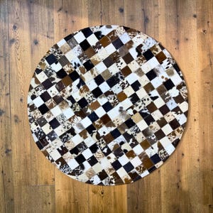 May include: A round cowhide rug with a patchwork design. The rug features a checkerboard pattern of black, white, brown, and tan squares. The rug is made of natural cowhide and has a soft, textured appearance.