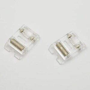 May include: Two clear plastic sewing machine presser feet, each featuring a metallic roller with a textured, cross-hatched surface. The components include small metal pins and adjustment screws integrated into the transparent molded plastic frame.