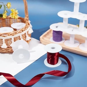 May include: A collection of white plastic spools, some empty and some wound with burgundy satin ribbon or purple yarn. The spools are arranged on a light wooden tray and inside a woven wicker basket decorated with white tassels and yellow flowers.