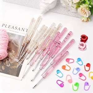 May include: A set of crochet hooks with clear handles containing gold or pink glitter, paired with a collection of colorful plastic stitch markers in red, pink, orange, yellow, green, blue, and purple, alongside a ball of pink yarn.