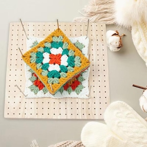 May include: A square wooden blocking board with a grid of holes and four metal pins holding two stacked crochet granny squares. The top square features a vibrant pattern of mustard yellow, teal, white, and orange yarn in a floral design.
