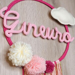 Puede incluir: Un adorno de pared circular rosa con el nombre "Eireuro" en hilo rosa. Cuenta con pompones blancos, rosas y fucsias, una nube blanca y borlas doradas y rosas. El aro está envuelto en hilo rosa.