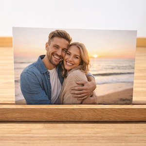 May include: A photograph of a smiling couple embracing on a beach at sunset, displayed in a wooden photo holder. The photo holder is made of light-coloured wood and sits on a wooden surface.