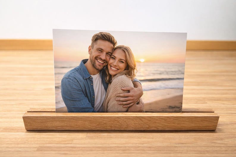 May include: A photograph of a smiling couple embracing on a beach at sunset, displayed in a wooden photo holder. The photo holder is made of light-coloured wood and sits on a wooden surface.