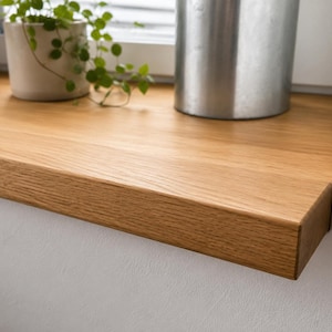 May include: A rectangular window sill made of light brown oak wood with a visible natural grain pattern. The sill is installed against a white wall and features a smooth, flat surface with a straight front edge and a clean, finished appearance.