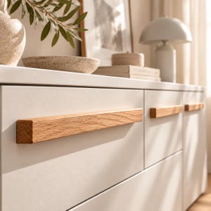 Könnte beinhalten: Ein weißer Schrank mit langen, horizontalen, rechteckigen Holzgriffleisten mit sichtbarer Maserung. Der Schrank hat eine glatte, matte Oberfläche und klare Linien, mit mehreren horizontal ausgerichteten Schubladen.