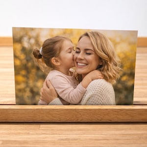 May include: A photograph of a mother and child, displayed in a wooden photo holder. The image shows a young girl kissing her mother on the cheek. The mother is smiling. The photo holder is made of light-coloured wood.