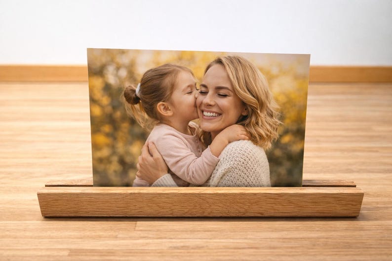 May include: A photograph of a mother and child, displayed in a wooden photo holder. The image shows a young girl kissing her mother on the cheek. The mother is smiling. The photo holder is made of light-coloured wood.