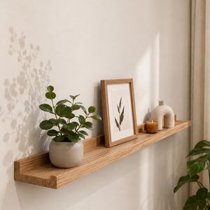 Könnte beinhalten: Ein langes Wandregal aus hellem Holz mit einer erhöhten Vorderkante. Auf dem Regal stehen ein kleiner grauer Keramiktopf mit einer grünen Pflanze, ein rechteckiger Holzbilderrahmen mit einem botanischen Druck, eine braune Kerze und eine beige Bogenfigur.