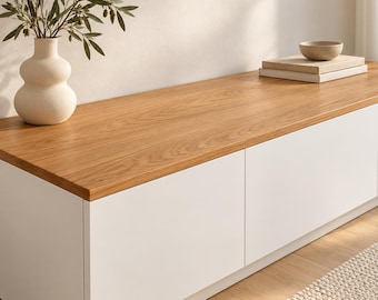 May include: A low rectangular storage bench with a smooth light brown wood grain top and solid white cabinet doors. On the left sits a matte cream-coloured ceramic vase with olive branches, and on the right are two stacked books with a small bowl.