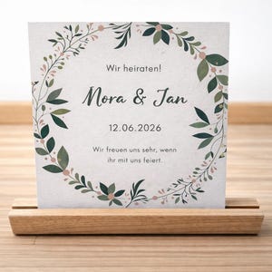 May include: A wedding invitation card with the names "Mora & Jan" and the date "12.06.2026" displayed on a light-colored background. The card is decorated with a floral wreath and is held in a wooden holder.