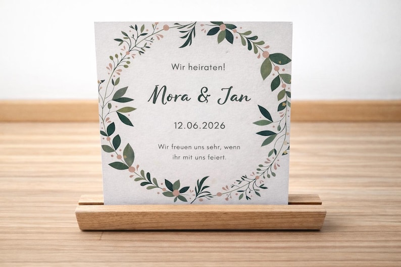 May include: A wedding invitation card with the names "Mora & Jan" and the date "12.06.2026" displayed on a light-colored background. The card is decorated with a floral wreath and is held in a wooden holder.