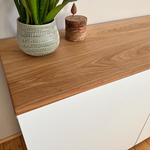 Könnte beinhalten: Ein weißer Schrank mit drei Türen und einer Holzplatte. Die Platte ist hellbraun mit einer Holzmaserung. Eine grüne Topfpflanze steht auf der Schrankplatte.