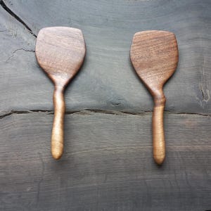 Puede incluir: Dos espátulas de madera con mangos marrones y veta de madera marrón claro. Las espátulas están hechas de madera de nogal y tienen un mango curvo.