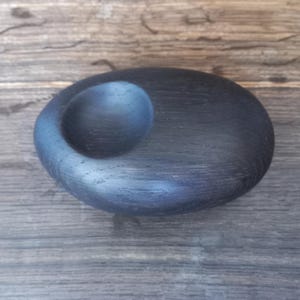 May include: A smooth, oval-shaped black wooden object with a single circular indentation on its top surface. The dark wood grain is visible across the rounded form, which features a polished finish and a solid, compact, and pebble-like appearance.