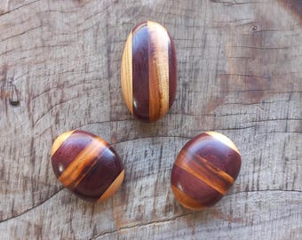 May include: Three wooden eggs with a striped pattern of brown, purple, and yellow wood. The eggs are on a wooden surface.