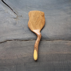 May include: A wooden spatula with a yellow handle. The spatula is curved and has a unique shape. It is made of natural wood and has a rustic look.