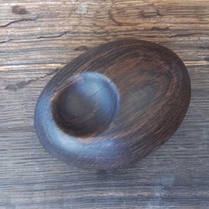 May include: A dark brown, oval-shaped wooden object with a smooth, polished finish and a single circular indentation centered on its top surface. The wood grain is visible throughout the piece, showing natural dark streaks and a deep brown tone.