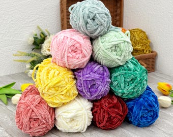 May include: A collection of colorful velvet yarn balls in shades of pink, yellow, purple, green, blue, and red. The yarn balls are stacked in a pyramid shape, with a wicker basket in the background.