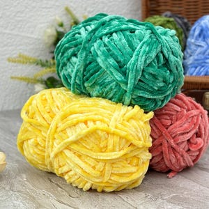 Può includere: Primo piano di diversi gomitoli di filato morbido e soffice in vari colori. Il filato è nelle tonalità del verde, del giallo e del rosso. Un cestino di vimini pieno di filato blu è sullo sfondo. Ideale per progetti di maglieria e uncinetto.