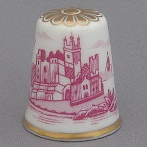 Puede incluir: Un dedal de porcelana blanca con un diseño rosa y dorado de un castillo. El dedal tiene una banda dorada alrededor de la parte superior e inferior.