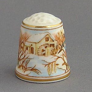 May include: A white porcelain thimble with a gold trim and a winter scene painted on it. The scene depicts a small house with snow on the roof and trees in the background.