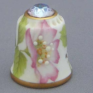May include: A decorative thimble with a floral design. The thimble features a pink and white flower with green leaves, accented with small white dots. A gold band encircles the top and bottom, and a faceted, clear gem adorns the top.