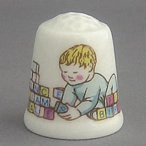 May include: A white porcelain thimble with a colorful illustration of a child playing with alphabet blocks. The child is wearing a blue shirt and has blonde hair. The blocks spell out the words "I AM ALL" and "BIRDS".