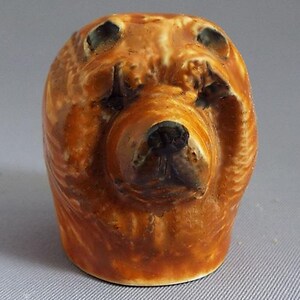 Puede incluir: Una figurita de cerámica de un perro marrón con ojos y nariz negros. El perro está sentado con la cabeza inclinada hacia abajo y las patas metidas debajo del cuerpo.