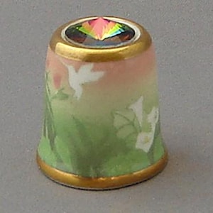 May include: A porcelain thimble with a green, pink, and white floral design. The thimble has a gold rim and a clear iridescent jewel on top.