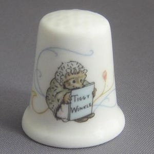 May include: A white ceramic thimble featuring a detailed illustration of a hedgehog character wearing a bonnet and shawl, holding a sign that reads Tiggy Winkle. The design includes delicate blue and orange swirling line accents.