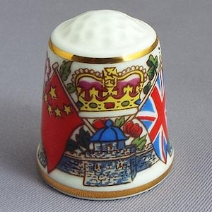 May include: A white porcelain thimble with a gold trim and a design featuring the flags of China and the United Kingdom, a crown, and a building.