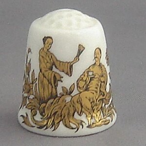 May include: White porcelain thimble with a gold illustration of two figures in a garden setting. One figure is holding a fan, and the other is seated, holding a cup. The thimble has a textured top.