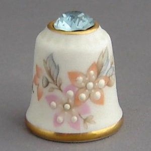 May include: A white porcelain thimble with a floral design and a light blue rhinestone on top. The thimble has a gold trim around the top and bottom.