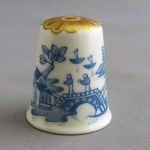 May include: A white ceramic thimble with a blue and gold design. The thimble features a scene of a bridge with people and boats, inspired by the classic blue and white willow pattern.